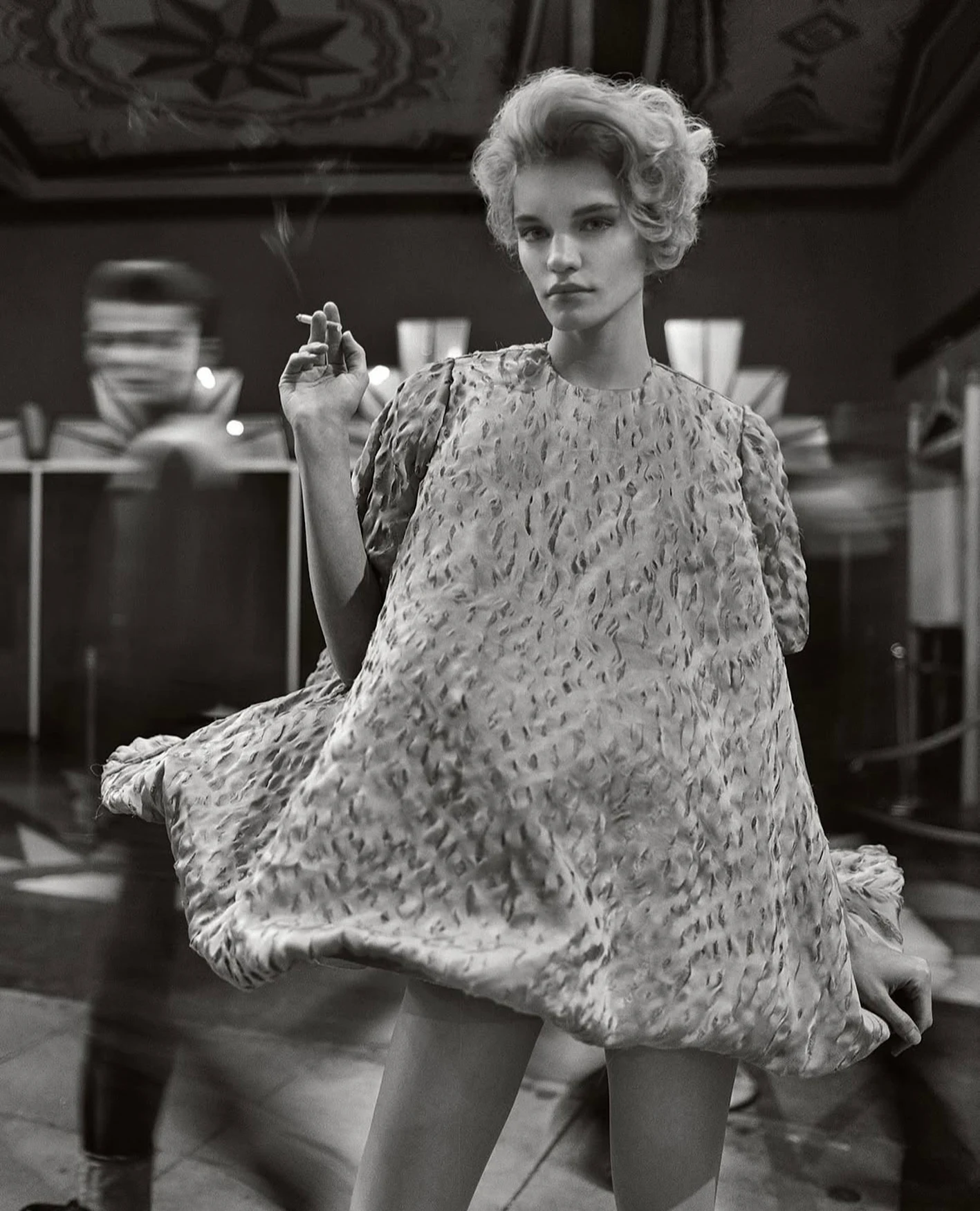 Monochrome portrait of a woman in a patterned dress holding a lit cigarette indoors, with blurred motion of a second person.