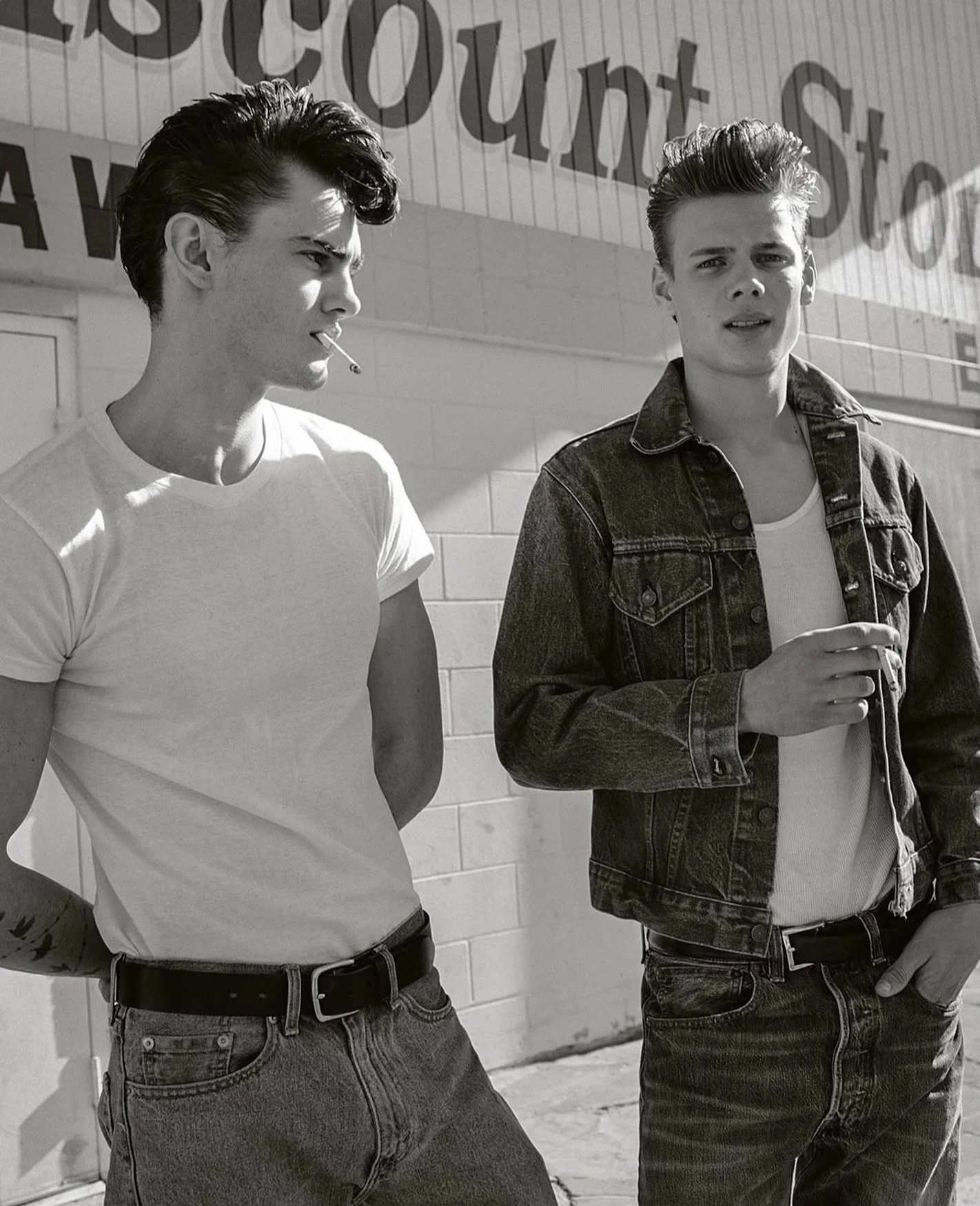 Two young adult men in 1950s style attire stand outside a discount store in a black and white medium shot.