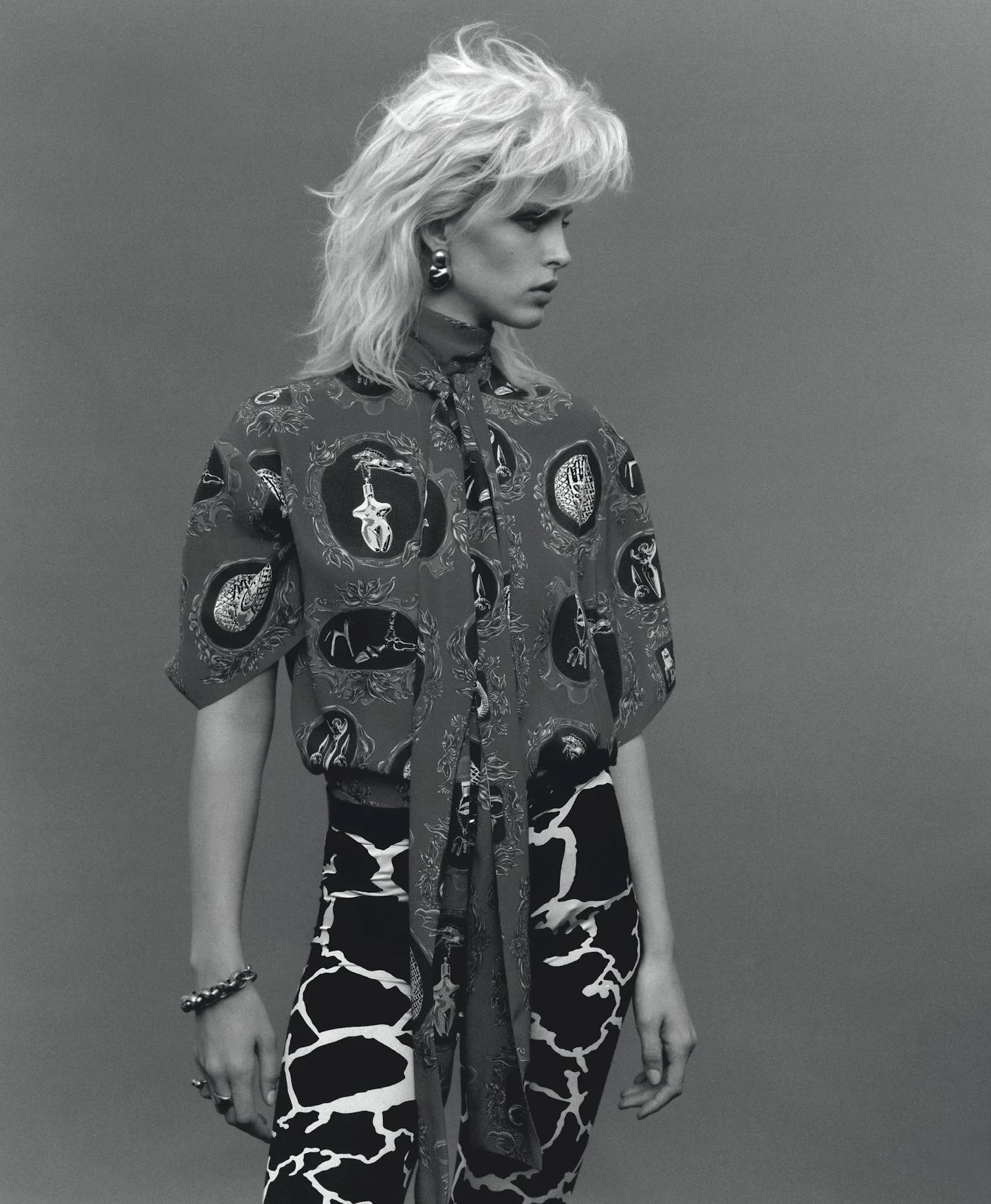 Woman with windswept blonde hair wearing patterned shirt and pants in studio portrait with grey backdrop. Fashion shot, medium angle.