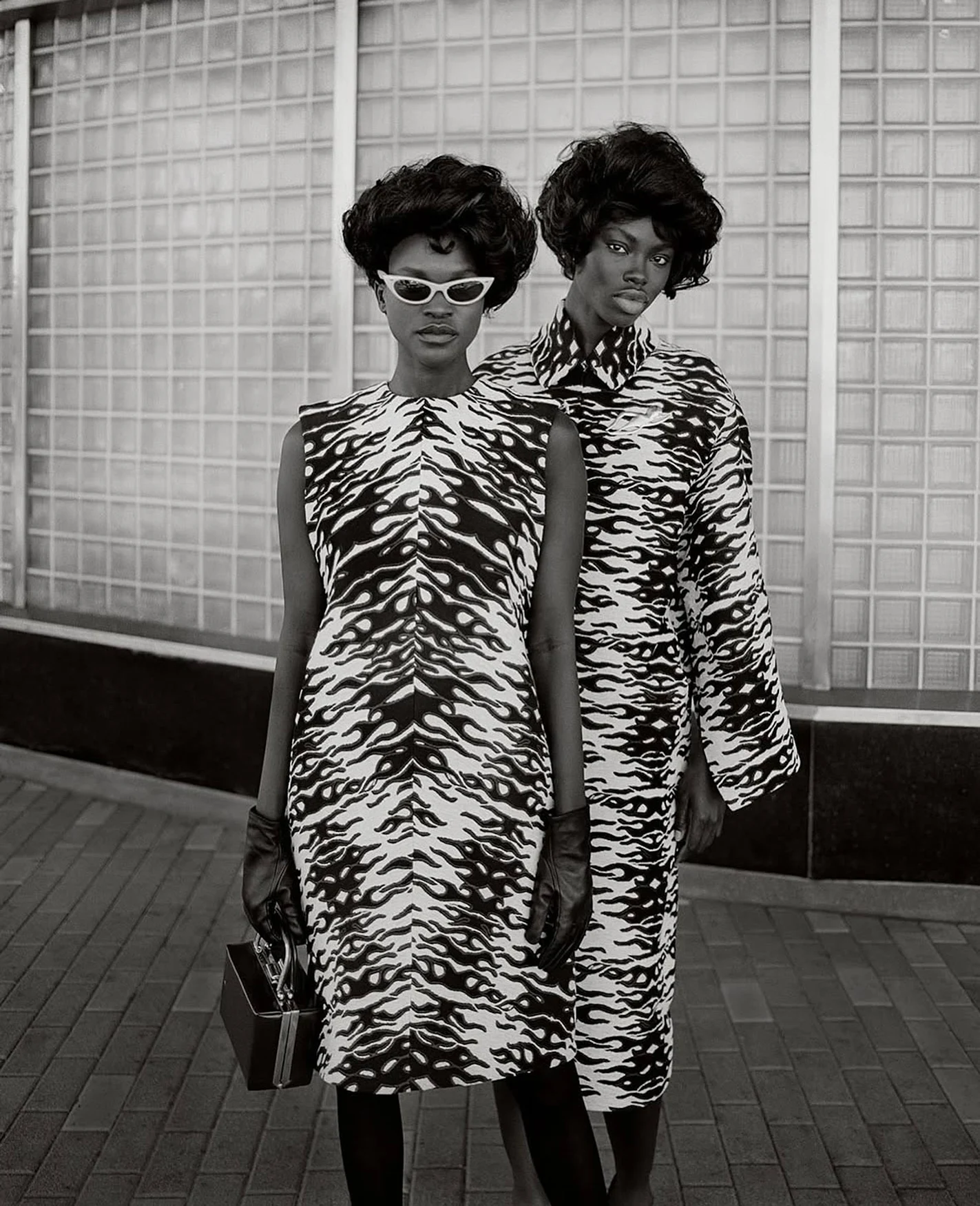 Monochrome fashion shot of two Black women in matching zebra-print dresses and retro hairstyles, standing against a textured glass block wall.