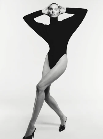 A woman with fair skin poses in a black long-sleeved bodysuit and black high heels against a white background in a studio setting.