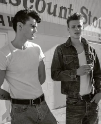 Two young men in white t-shirts and denim standing outdoors in front of a light-colored brick wall.
