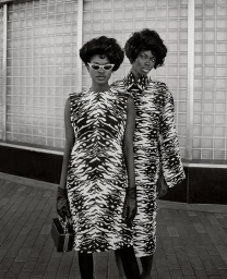 Two black female models in patterned dresses and dark glasses standing outdoors with structured hairstyles and gloves