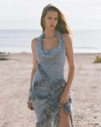 Model with long blonde hair wearing a textured blue halter dress standing on a sandy beach