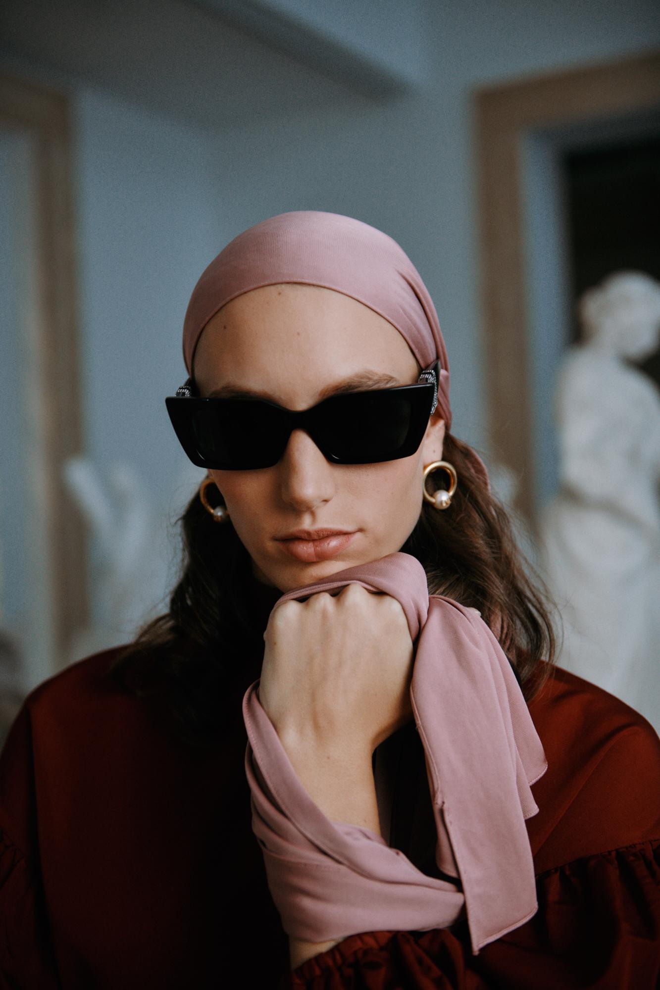 Portrait of a stylish adult woman wearing a headscarf and sunglasses, posed against a neutral background with classical sculpture.