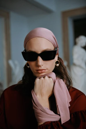 Portrait of a stylish adult woman wearing a headscarf and sunglasses, posed against a neutral background with classical sculpture.