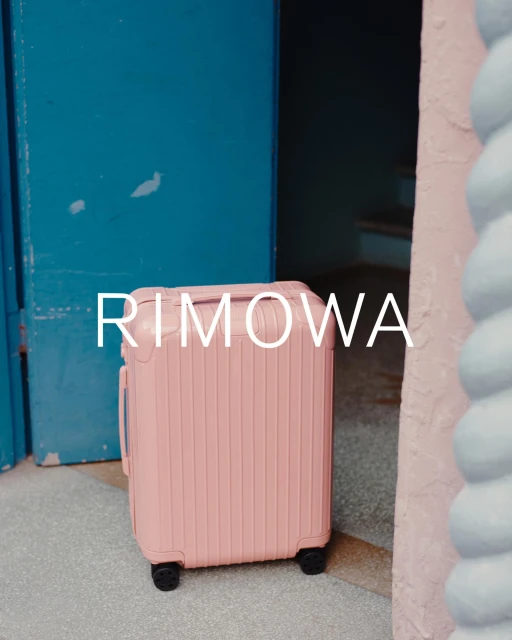 Coral pink Rimowa suitcase positioned in front of a turquoise door and a light pink wall, creating a vibrant and modern travel image.