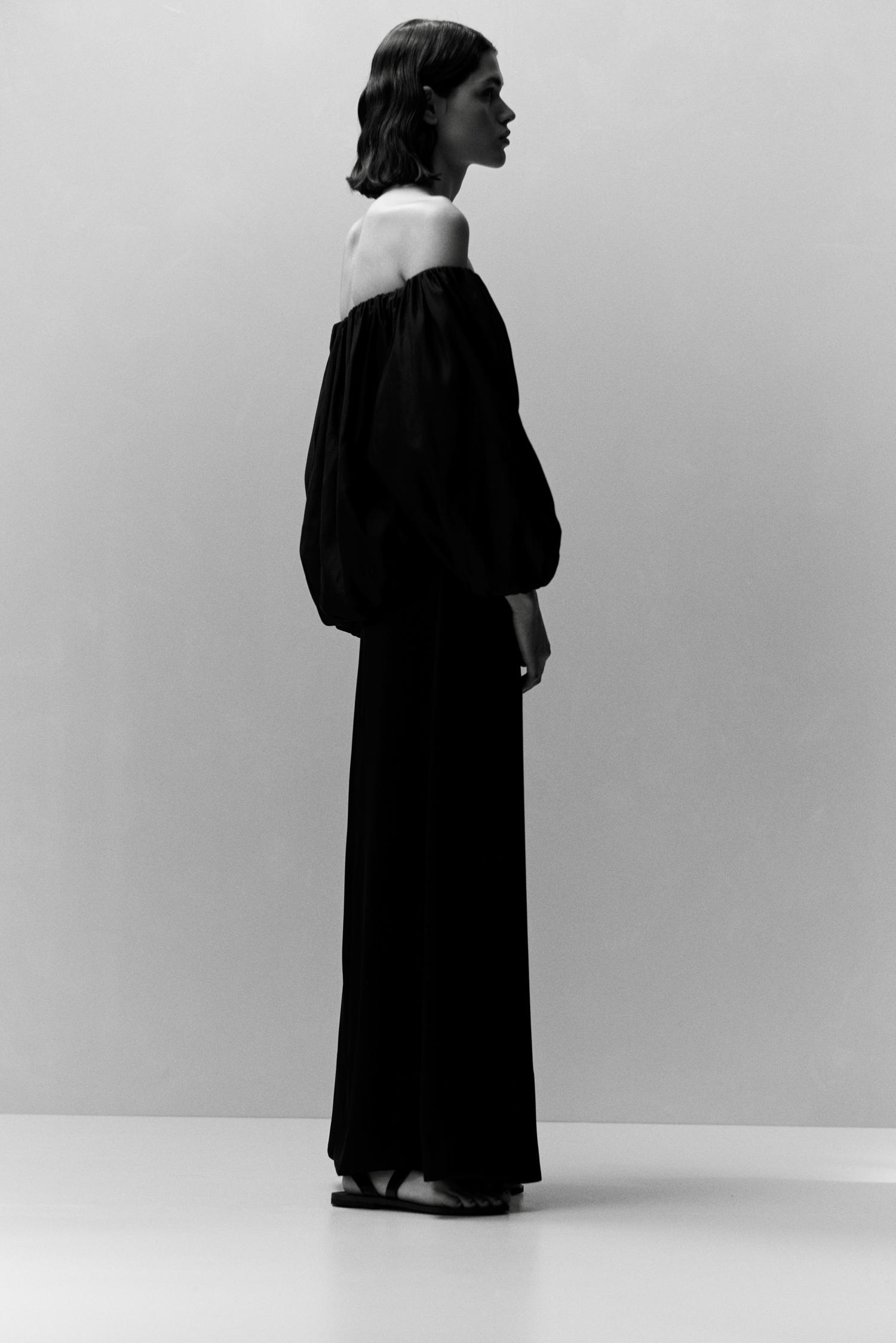 A woman in a black off-the-shoulder dress standing in a photo studio with a neutral background, captured in a medium shot.