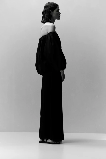 A woman in a black off-the-shoulder dress standing in a photo studio with a neutral background, captured in a medium shot.