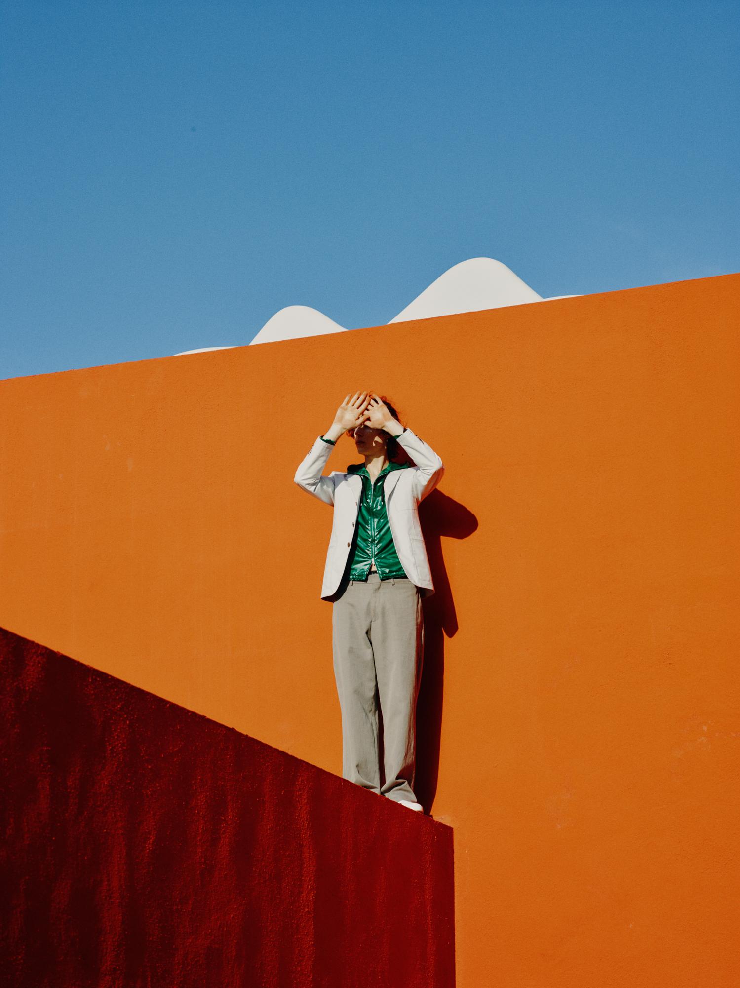 Fashionable person poses in front of an orange wall in bright sunlight, wearing a suit and covering their face with hands.