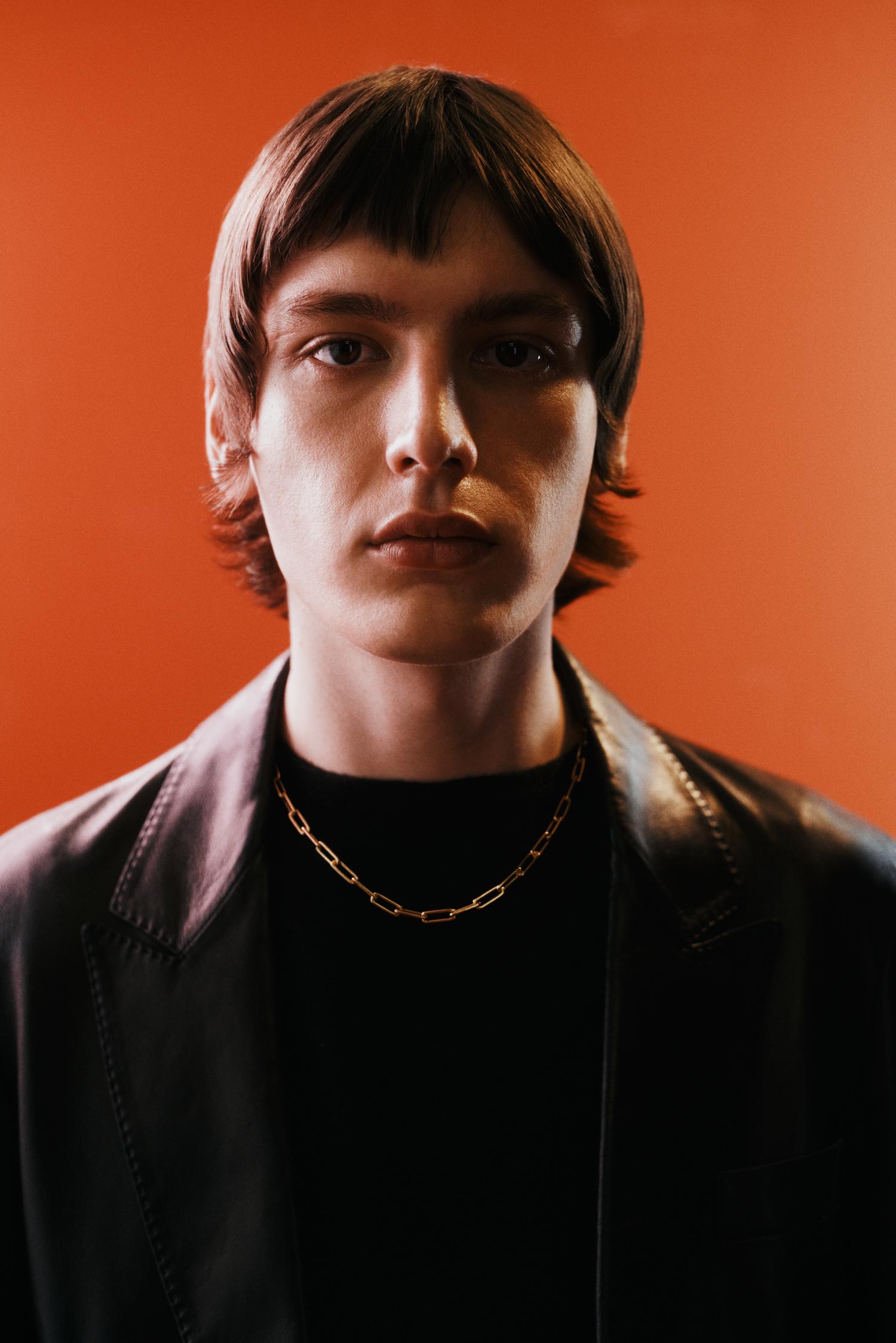 Portrait of a dark-haired male wearing a gold chain and black jacket against an orange background, focusing on fashion and portraiture.