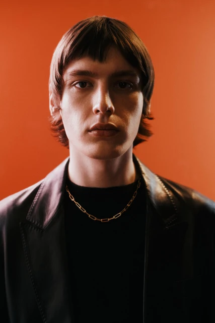 Portrait of a dark-haired male wearing a gold chain and black jacket against an orange background, focusing on fashion and portraiture.