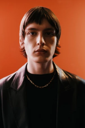 Portrait of a dark-haired male wearing a gold chain and black jacket against an orange background, focusing on fashion and portraiture.