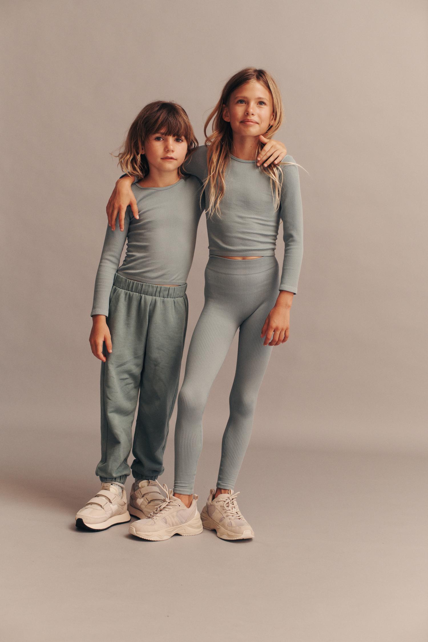 Two young children stand in a studio, one with an arm around the other, wearing matching ribbed tops, leggings, and sweatpants with sneakers.