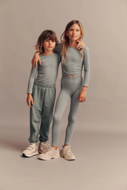 Two young children stand in a studio, one with an arm around the other, wearing matching ribbed tops, leggings, and sweatpants with sneakers.