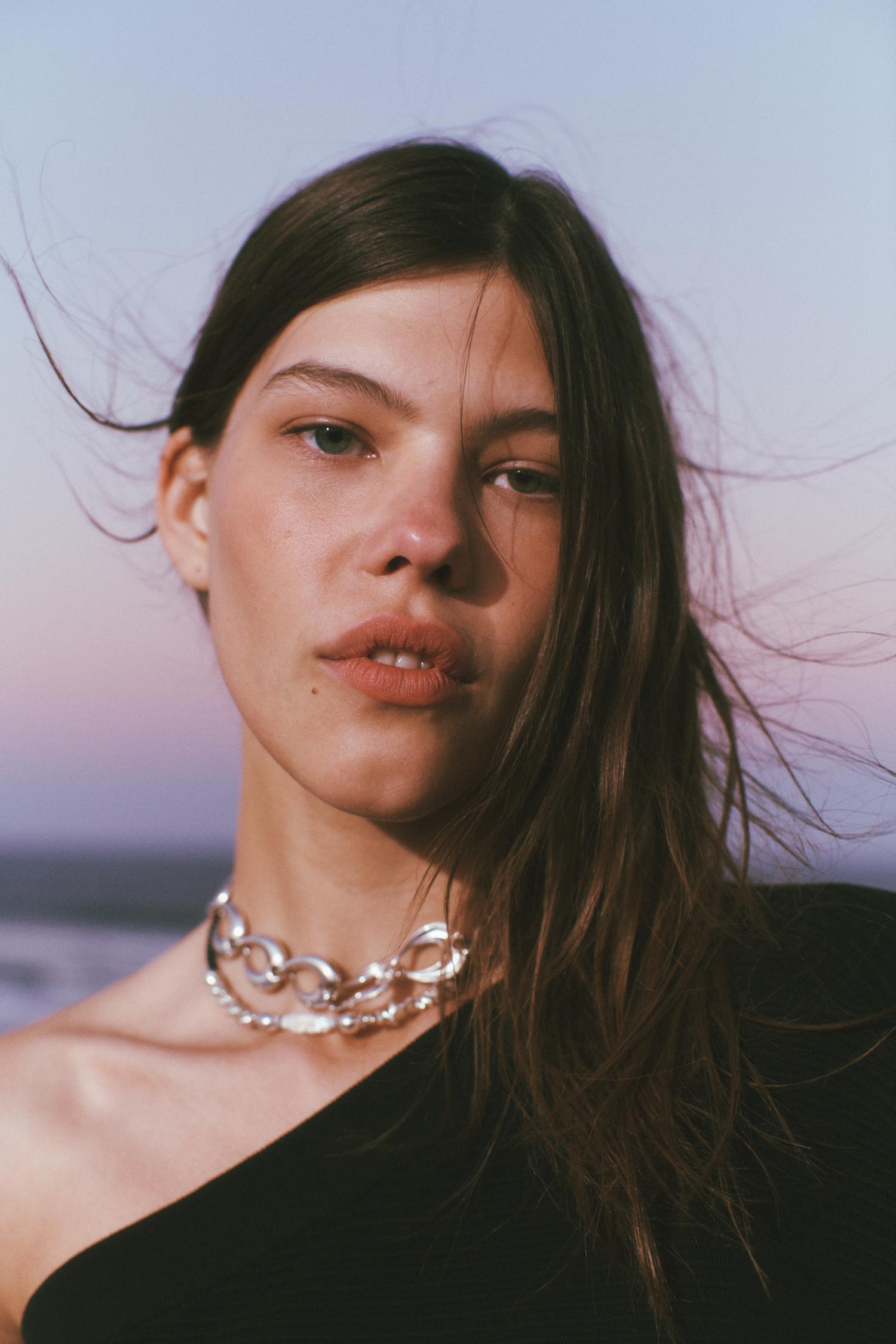 Close-up of a young adult woman wearing a silver chain necklace, shot against a soft, graduated blue to purple background.