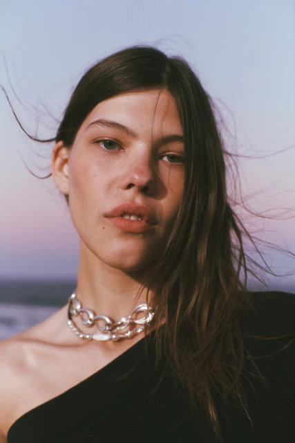 Close-up of a young adult woman wearing a silver chain necklace, shot against a soft, graduated blue to purple background.