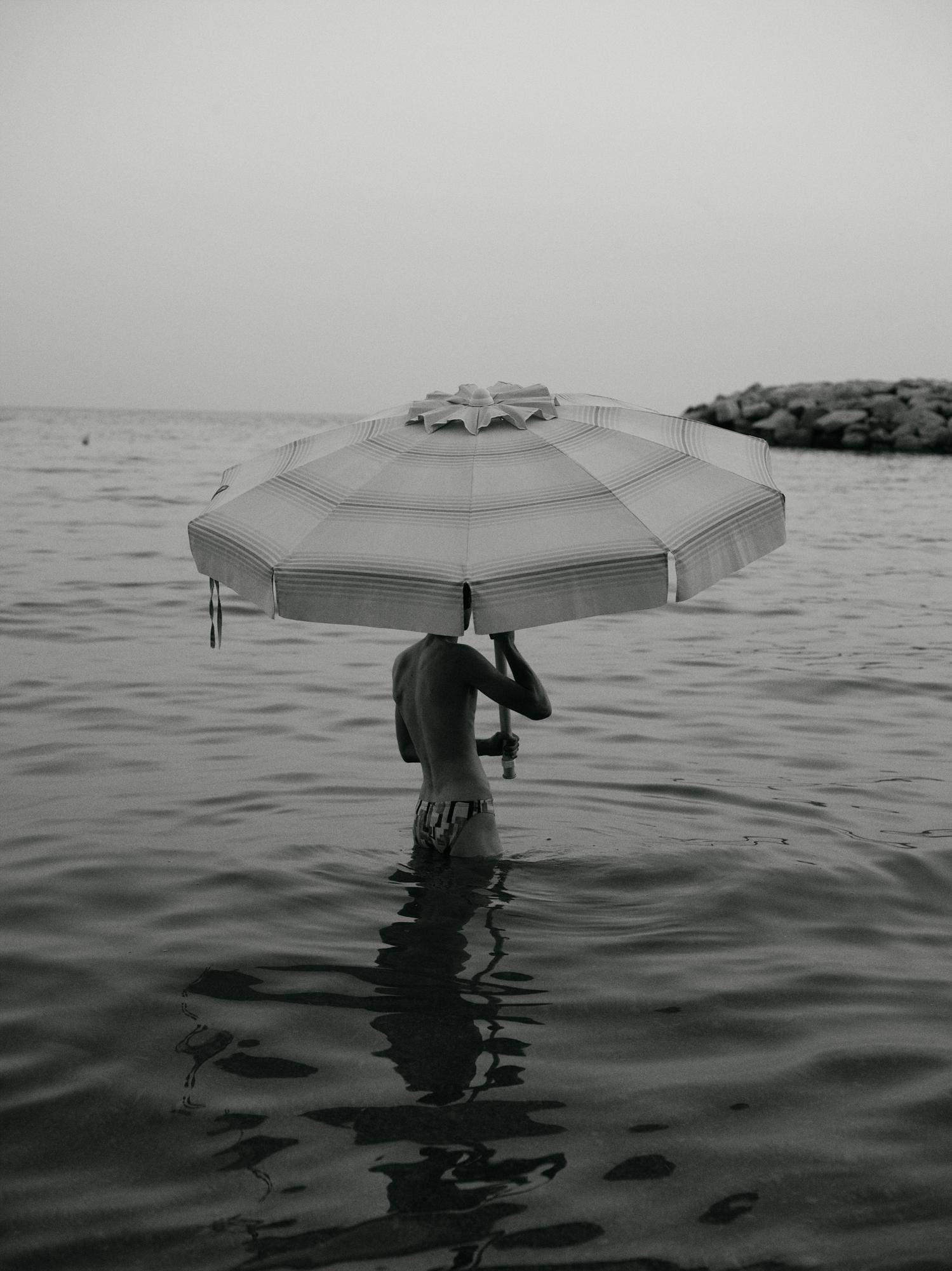 Monochromatic image of a child standing in ocean water holding a striped beach umbrella over their head near a rocky shoreline.