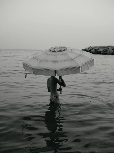 Monochromatic image of a child standing in ocean water holding a striped beach umbrella over their head near a rocky shoreline.