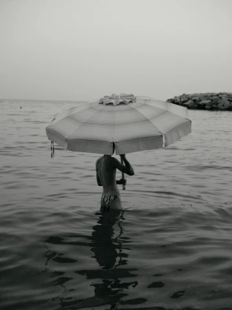 Monochromatic image of a child standing in ocean water holding a striped beach umbrella over their head near a rocky shoreline.