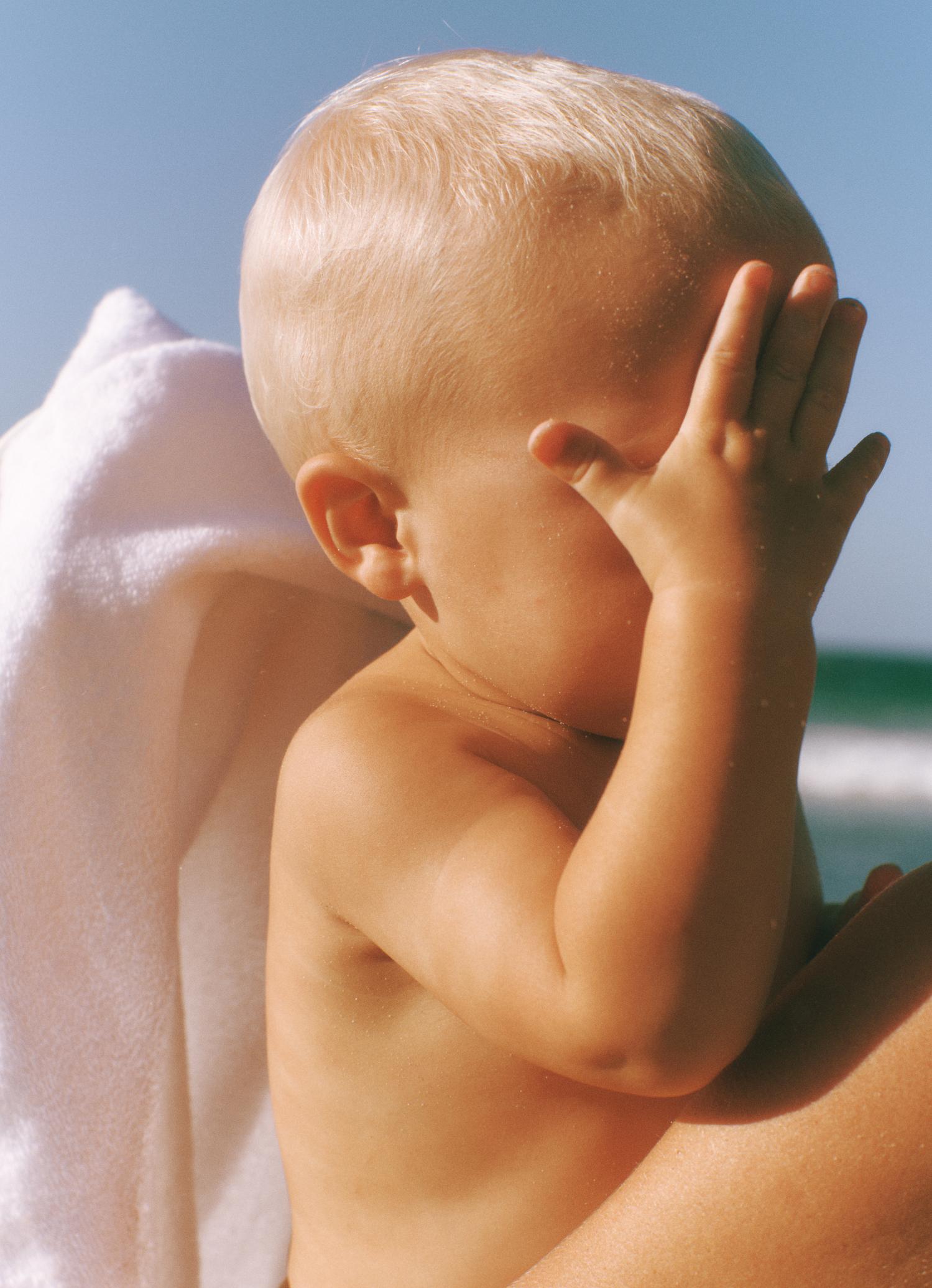 A young child with fair hair sits outside, shielding their face with a hand near a white towel. The ocean and blue sky are in the background.