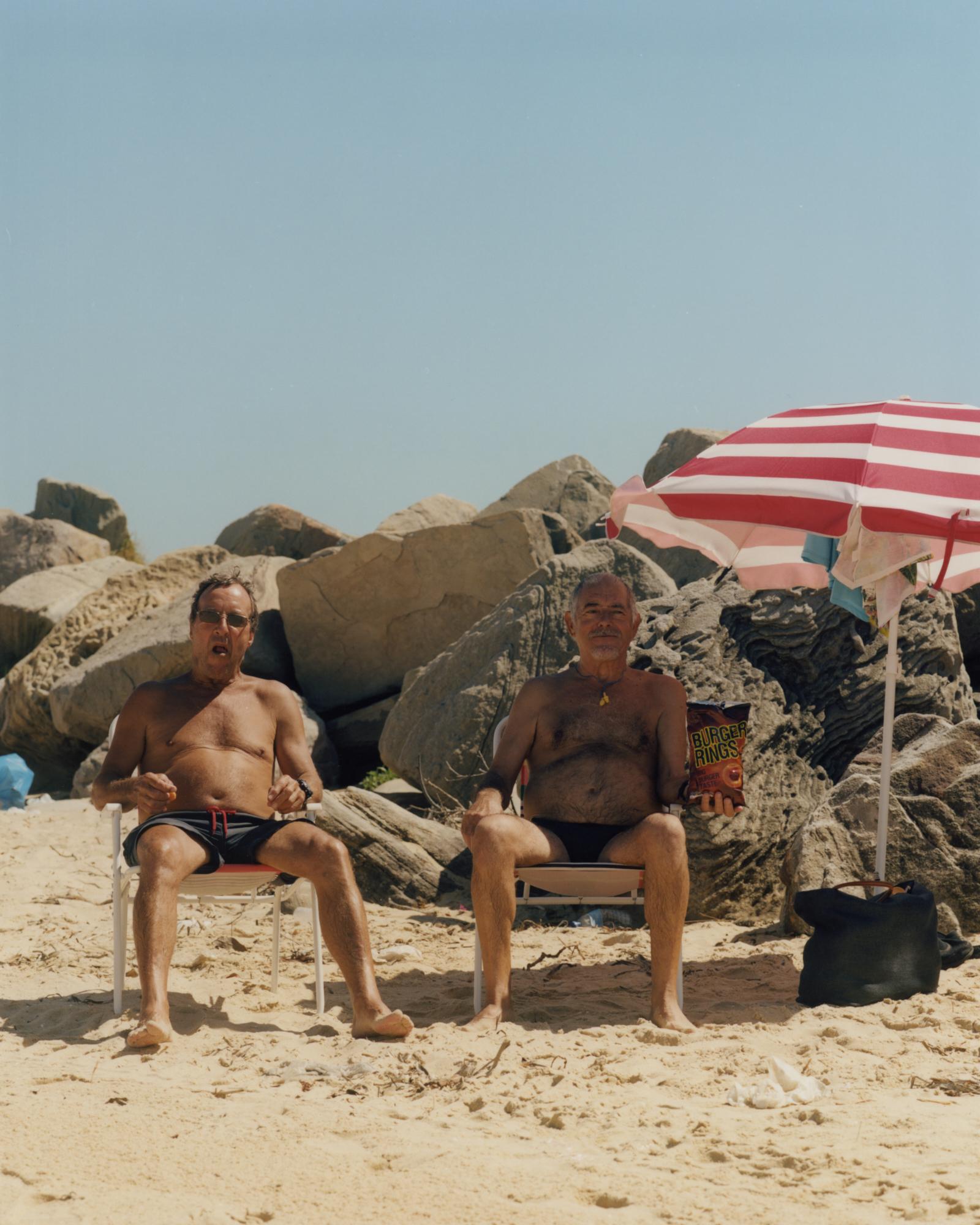 Two shirtless men sitting on beach chairs under an umbrella, relaxing on a sandy beach with rocks in the background on a sunny day.