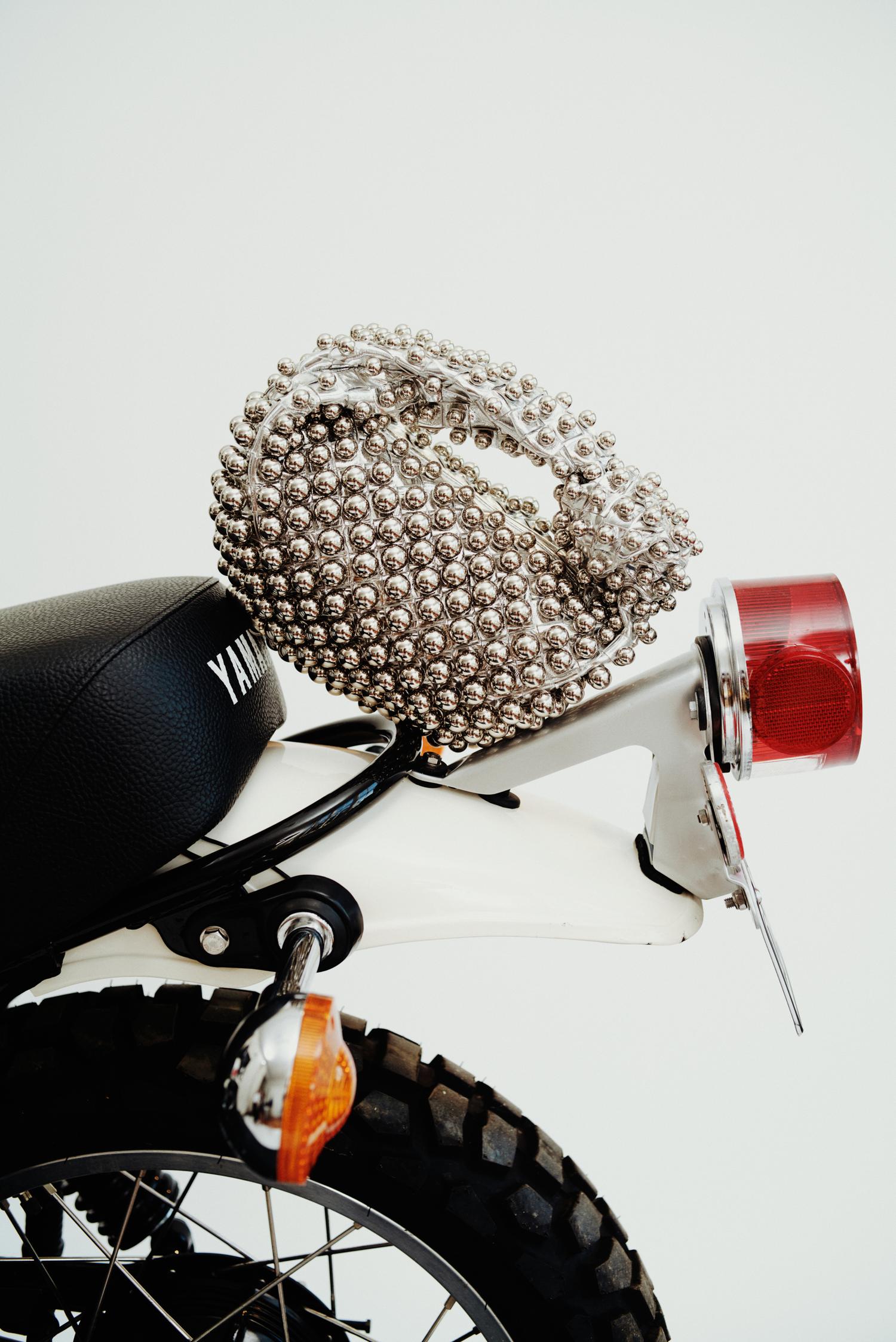 Rear view of a vintage Yamaha motorcycle with a black seat, white fender, and silver handbag covered in silver baubles on the seat.