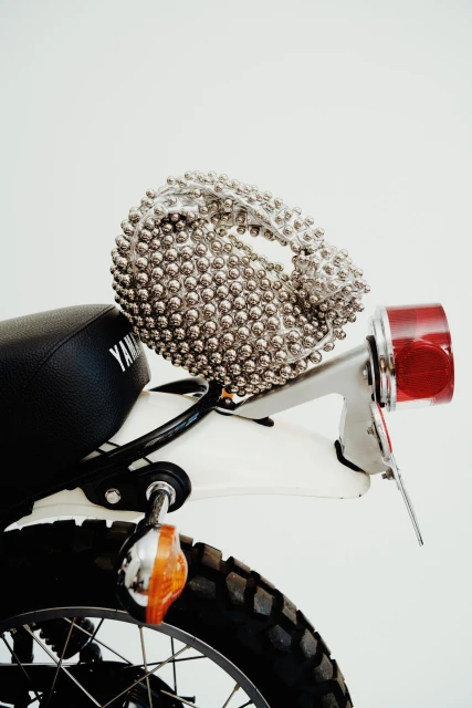 Rear view of a vintage Yamaha motorcycle with a black seat, white fender, and silver handbag covered in silver baubles on the seat.