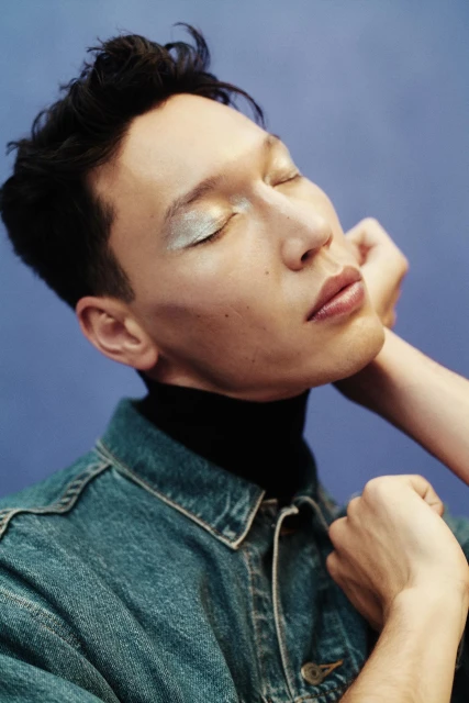 Close-up portrait of a person with light skin, dark hair, wearing a denim jacket and shimmering makeup, posing against a blue background.