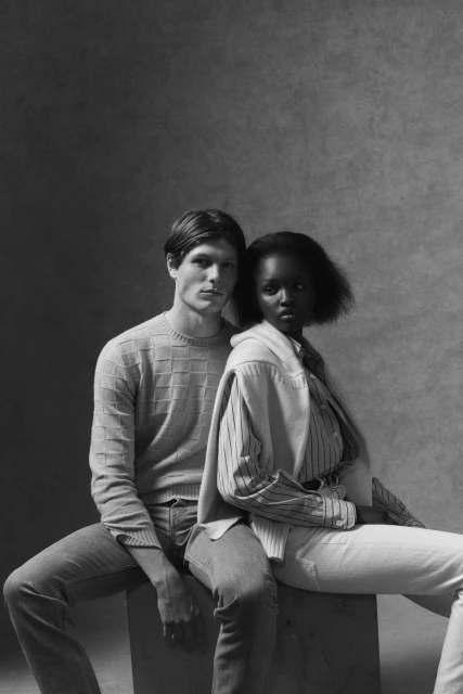 Monochrome portrait of a light-skinned male and dark-skinned female fashion models in casual wear sitting on a box in a studio.
