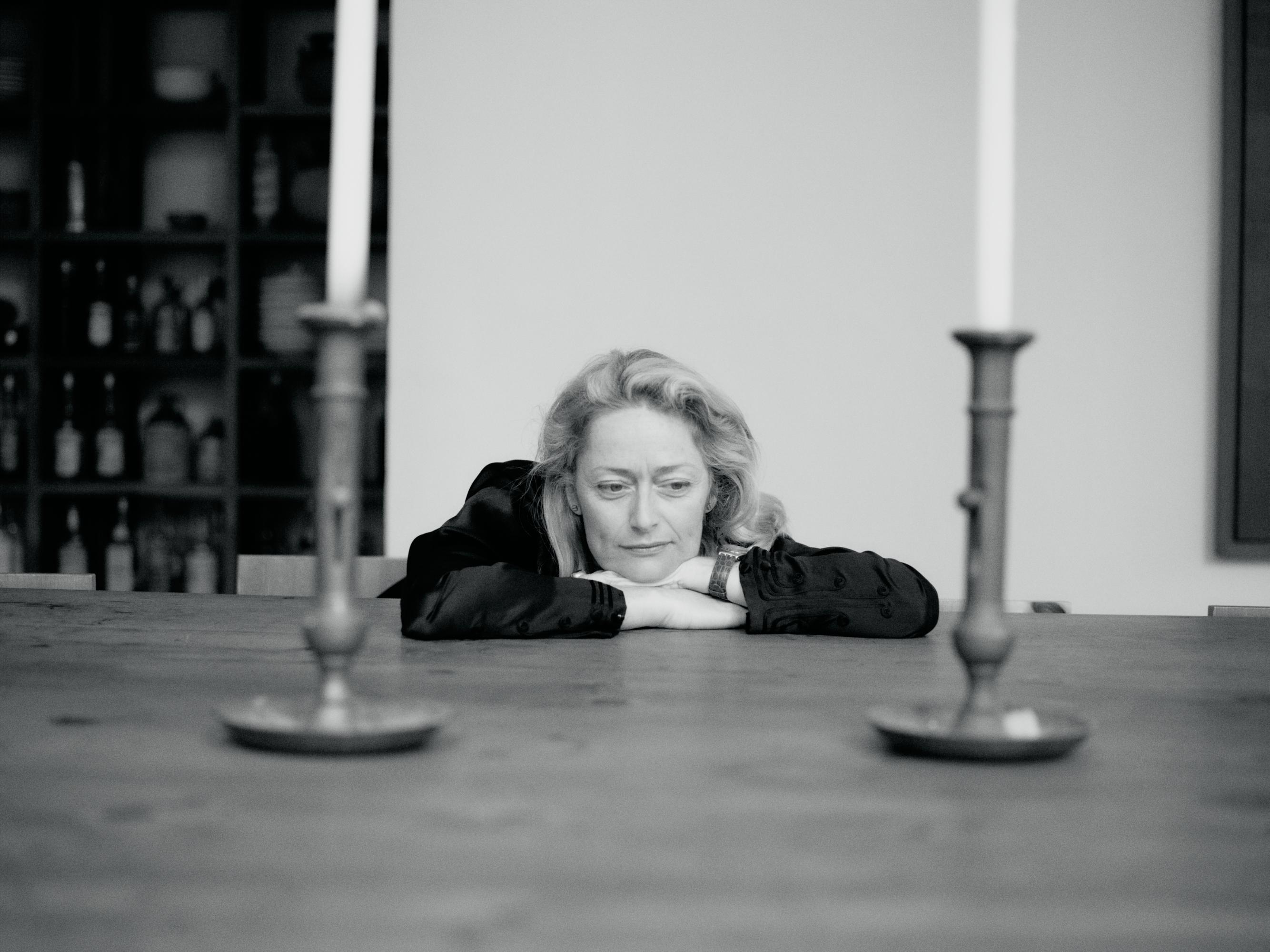 Black and white portrait of an adult woman resting her head on her arms at a wooden table, with two candlesticks in the foreground.