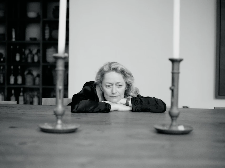 Black and white portrait of an adult woman resting her head on her arms at a wooden table, with two candlesticks in the foreground.