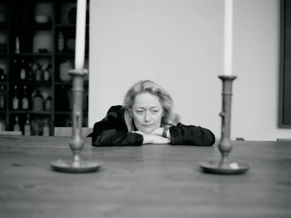 Black and white portrait of an adult woman resting her head on her arms at a wooden table, with two candlesticks in the foreground.