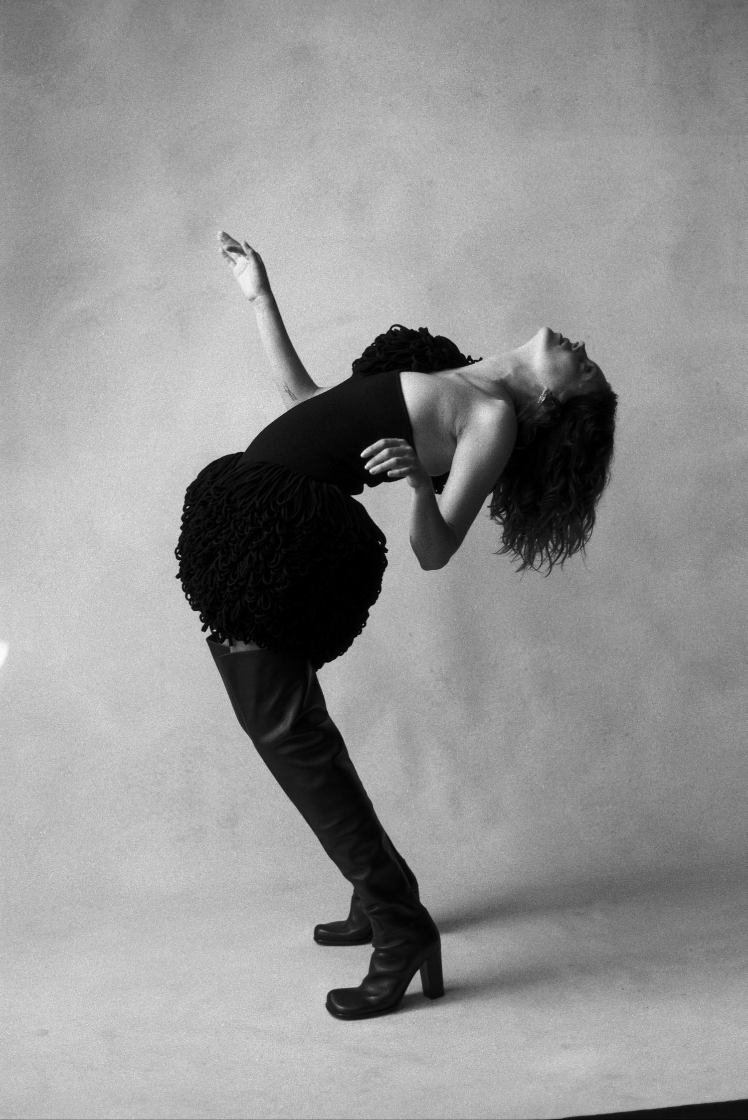 Full-length portrait of a woman in a black dress and thigh-high boots leaning back against a neutral background in a studio setting.