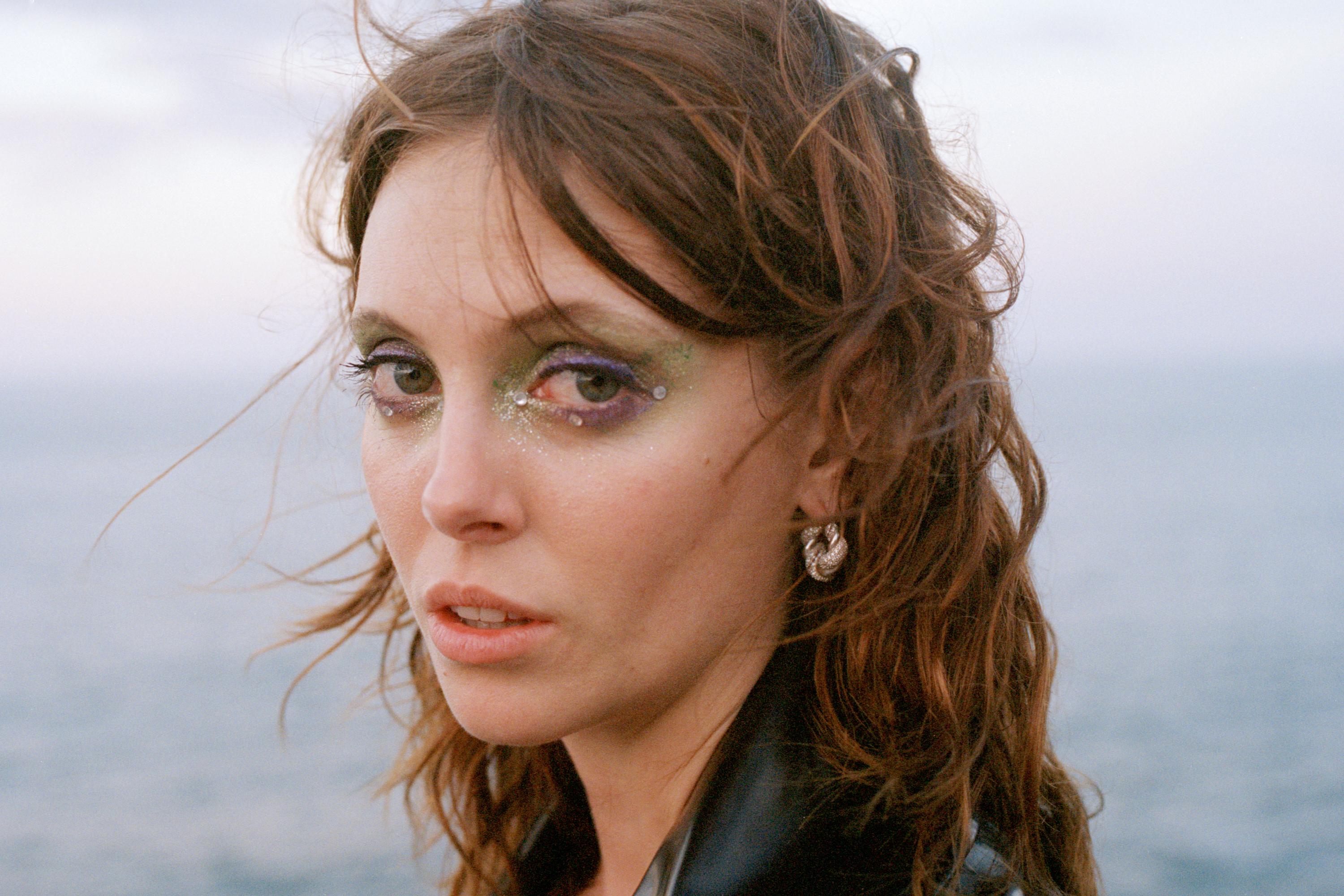 Close-up portrait of a young woman with creative eye makeup posing outdoors against a blurred backdrop of water and overcast sky.
