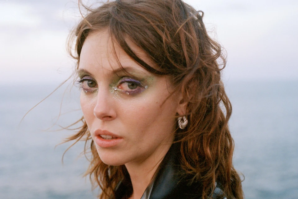 Close-up portrait of a young woman with creative eye makeup posing outdoors against a blurred backdrop of water and overcast sky.