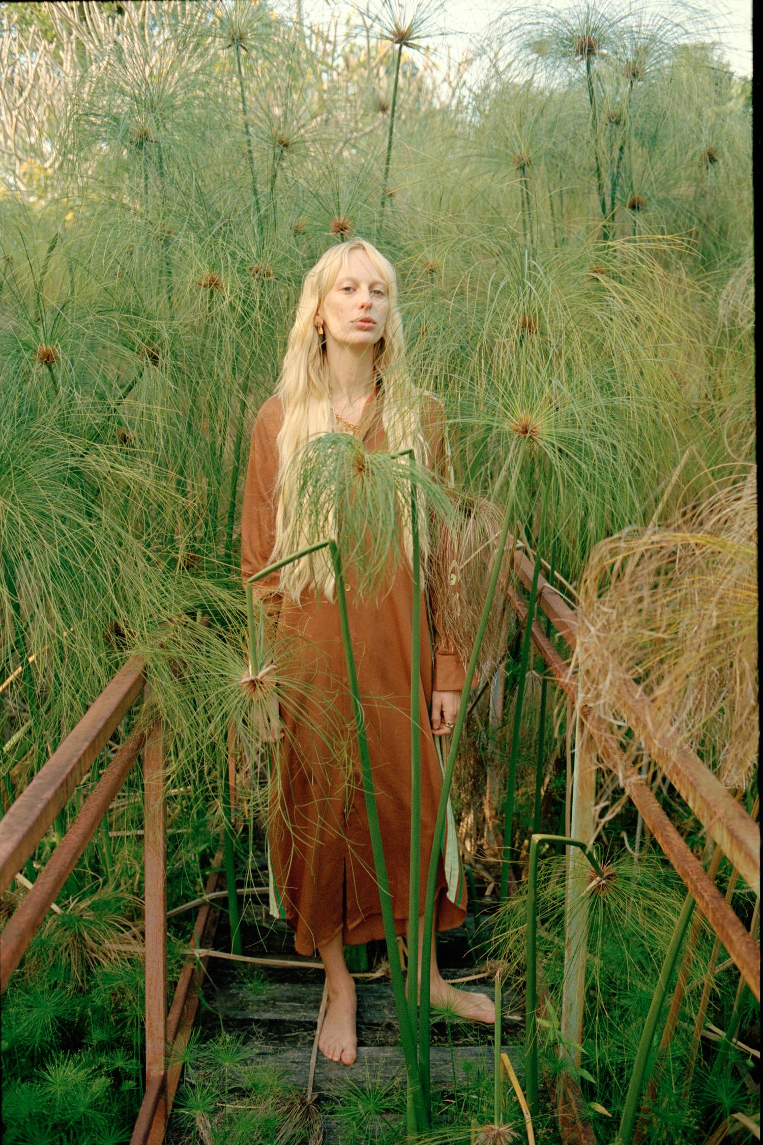 Blonde woman stands on a wooden walkway amidst lush green vegetation, wearing a brown dress, in a natural, outdoor setting with warm lighting.