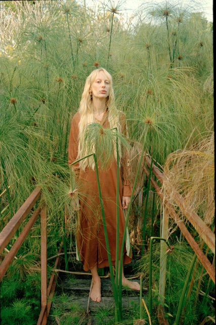 Blonde woman stands on a wooden walkway amidst lush green vegetation, wearing a brown dress, in a natural, outdoor setting with warm lighting.