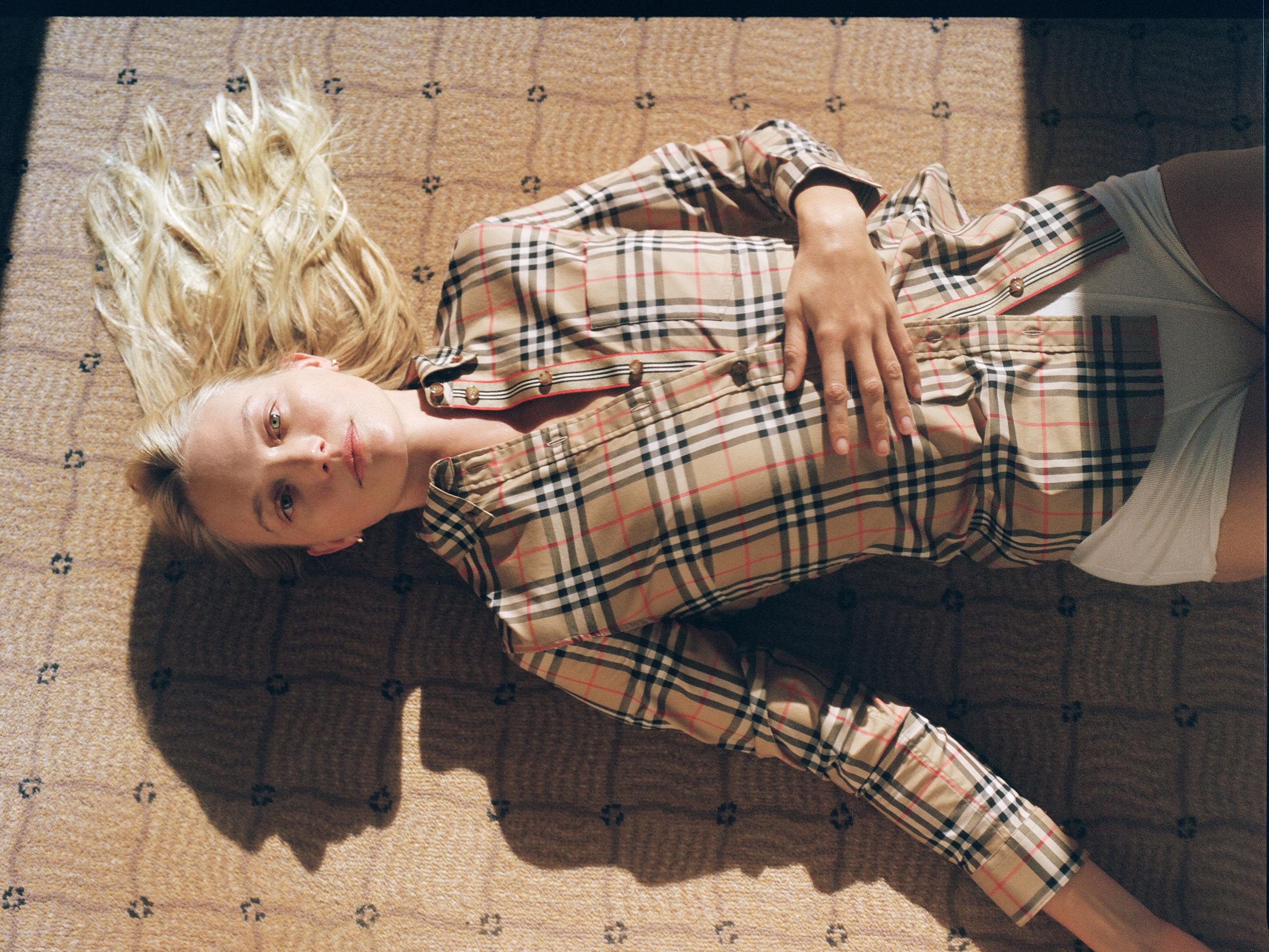 A blond female model poses on a patterned carpet indoors wearing a Burberry-patterned shirt and white shorts in natural light.