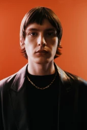 Portrait of a man with dark hair wearing a black jacket and gold chain necklace against an orange background