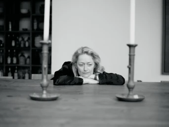 Woman resting her chin on her crossed arms at a wooden table in a room, framed by two tall candlesticks.