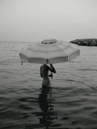 A person standing in the water holding a large beach umbrella over their head against a flat horizon.