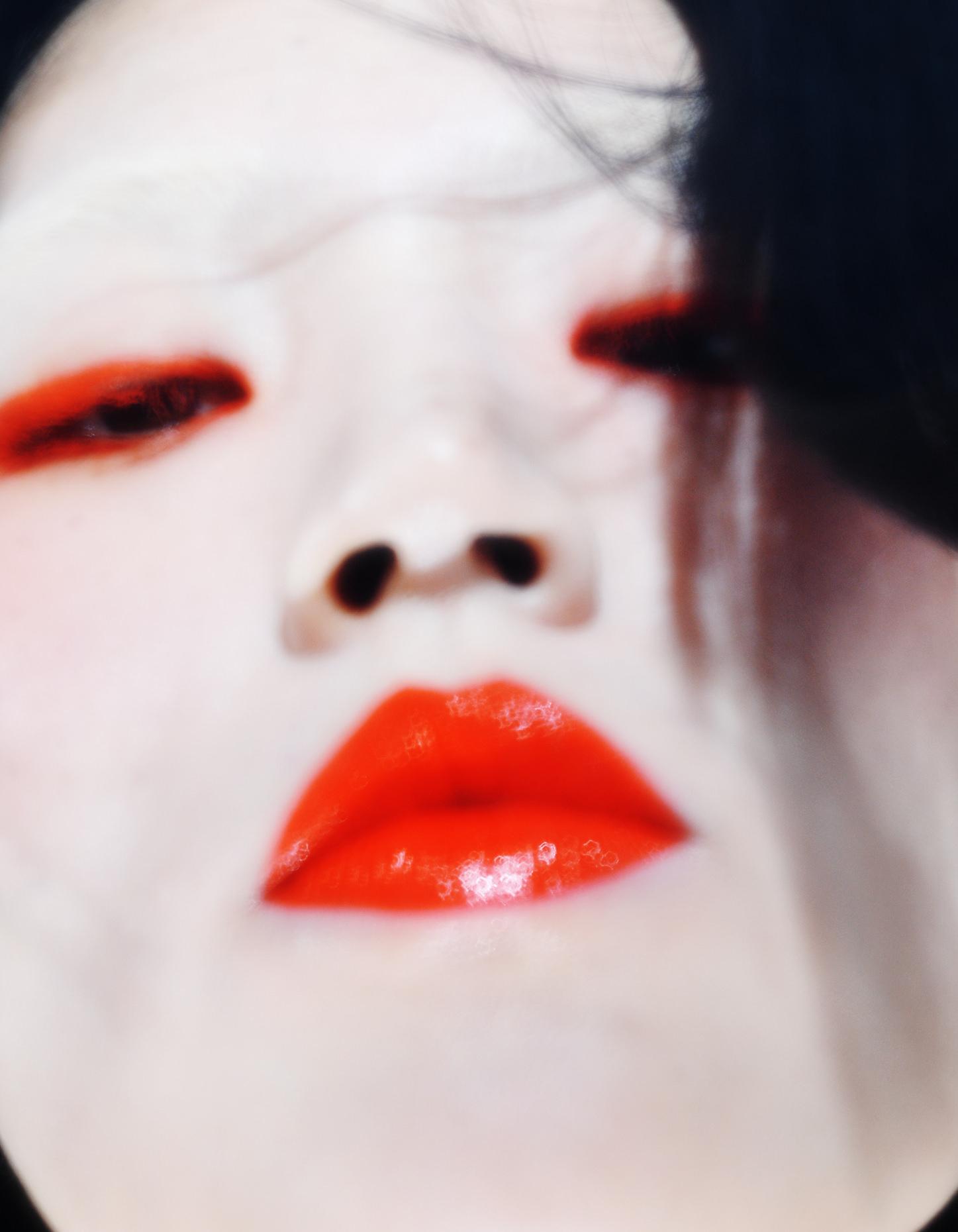 Close-up portrait of an adult female with bright red glossy lipstick and matching red eye makeup on light skin against a dark background.
