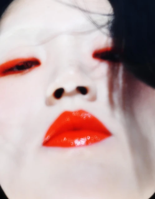 Close-up portrait of an adult female with bright red glossy lipstick and matching red eye makeup on light skin against a dark background.