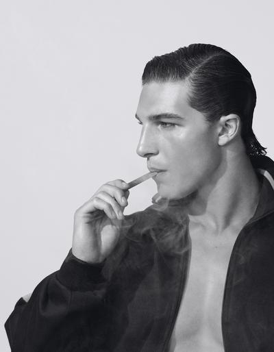 Monochrome close-up of an adult male in a jacket, smoking, and looking off to the side in a brightly lit, indoor studio environment.