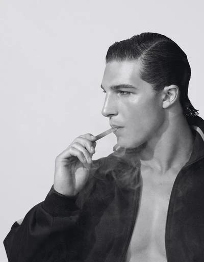 Monochrome close-up of an adult male in a jacket, smoking, and looking off to the side in a brightly lit, indoor studio environment.
