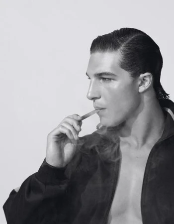 Monochrome close-up of an adult male in a jacket, smoking, and looking off to the side in a brightly lit, indoor studio environment.