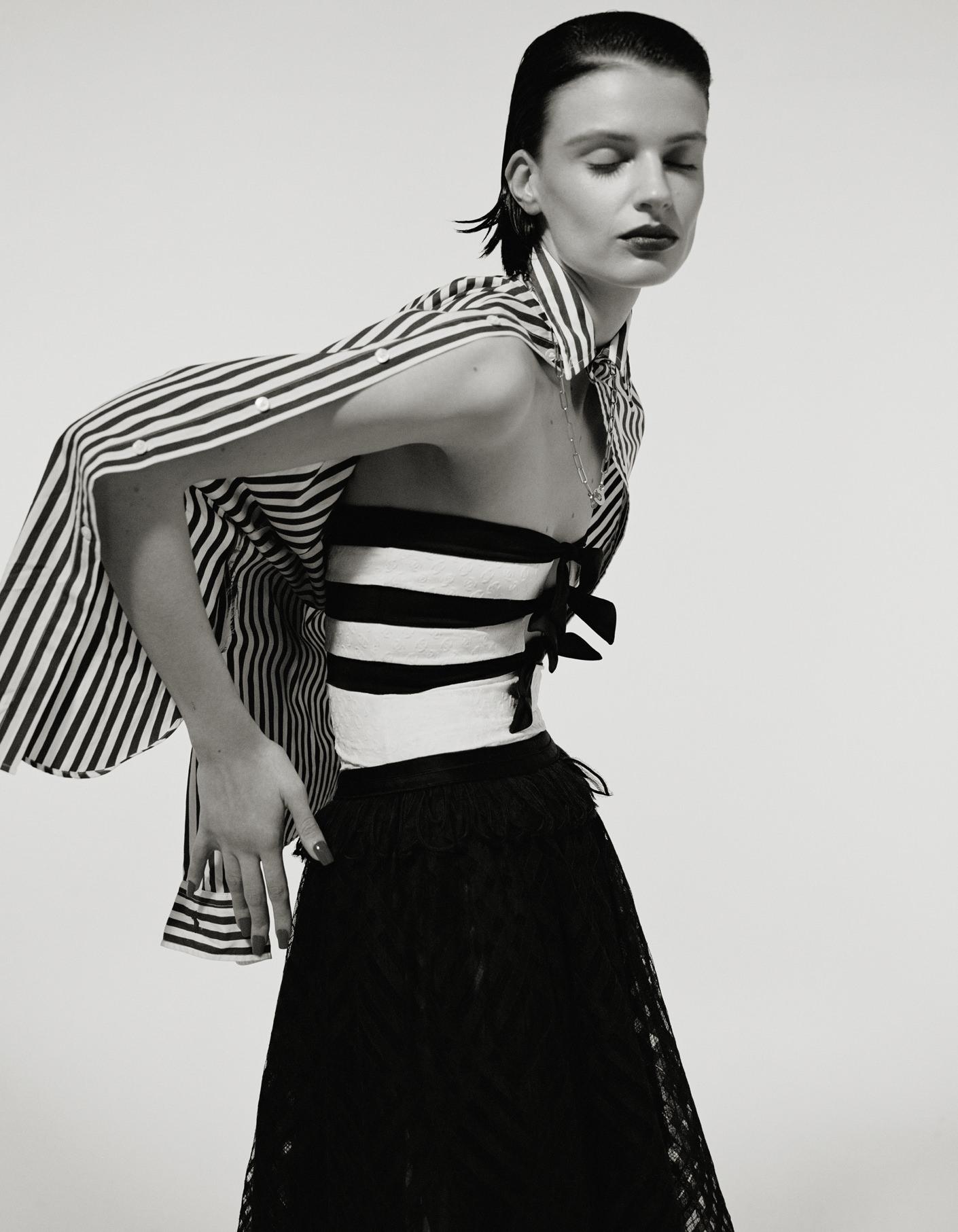 Monochrome portrait of a woman in a striped top and lace skirt, posing in front of a white backdrop in a fashion editorial style.