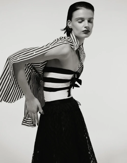 Monochrome portrait of a woman in a striped top and lace skirt, posing in front of a white backdrop in a fashion editorial style.