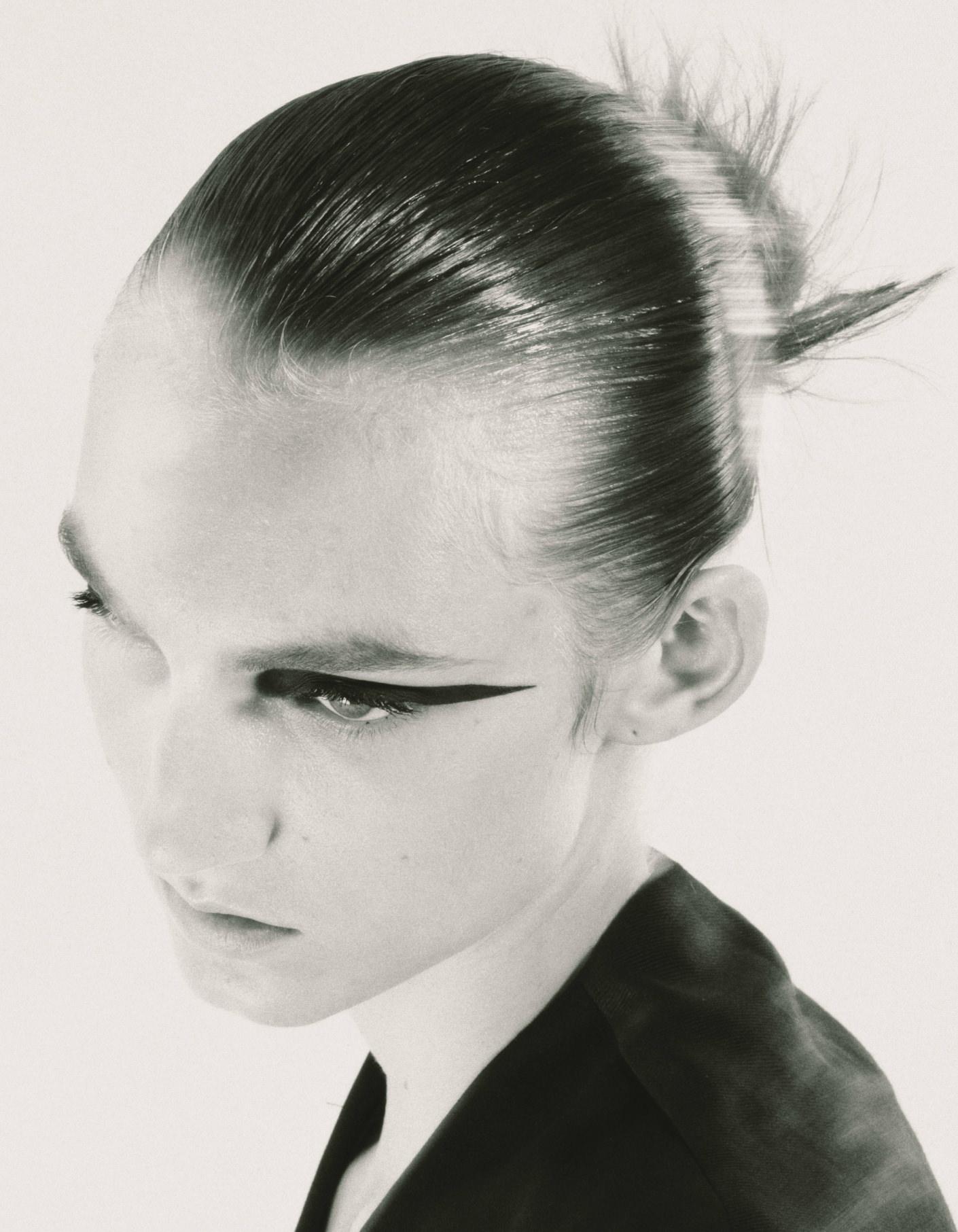 Close-up portrait of a model with sleek pulled-back hair and dramatic winged eyeliner against a plain background.